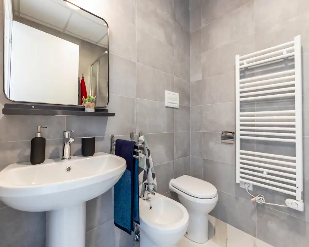 Bagno moderno e ben illuminato con lavabo a colonna, bidet e WC, ampia specchiera e scalda salviette; piastrelle grigie neutre e accessori ordinati.