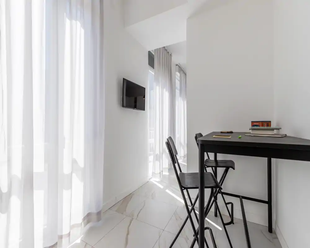 Zona giorno luminosa e minimal con tende leggere a tutta altezza, TV a muro e un piccolo tavolo alto con sedie — ideale per pasti veloci o lavoro vicino alla finestra.