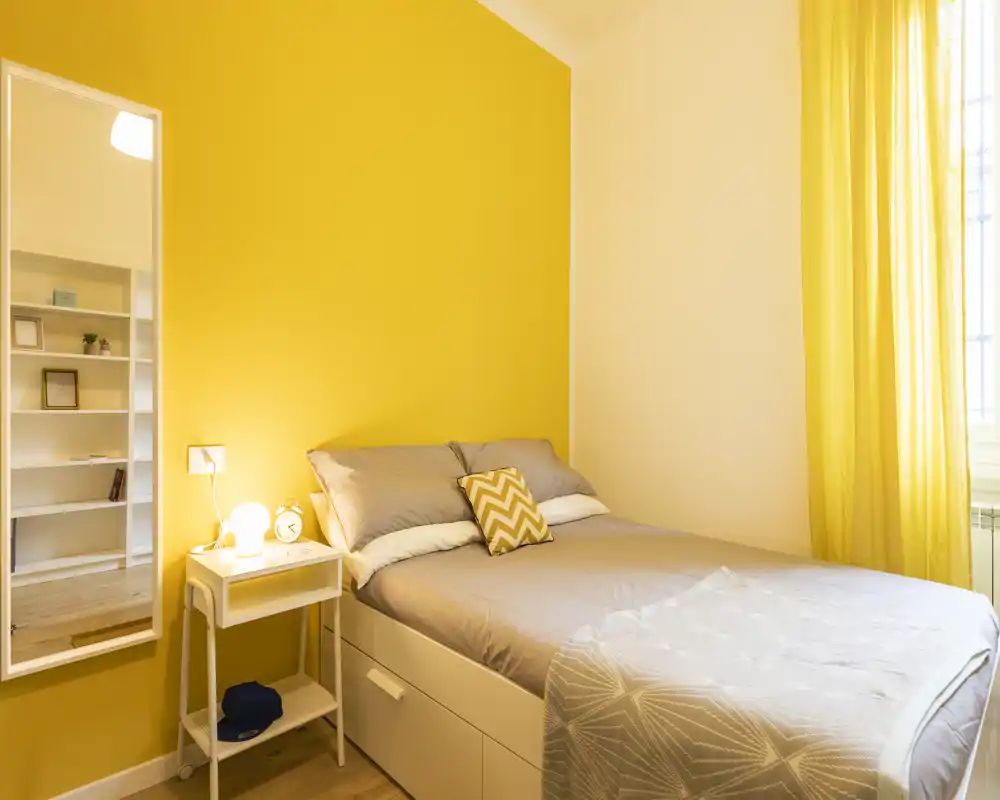 Chambre compacte et lumineuse avec un lit double confortable, un mur jaune chaleureux et des rideaux assortis. La table de chevet avec lampe et miroir offre un espace accueillant, parfait pour la location.