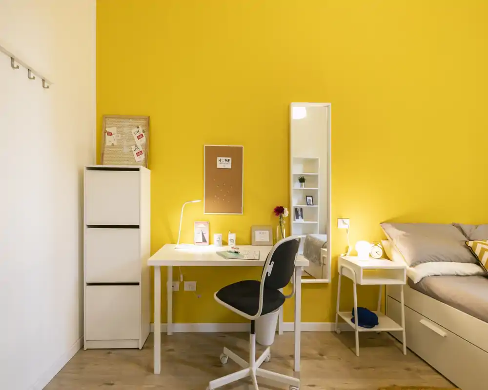 Coin chambre lumineux et moderne avec bureau blanc et chaise devant un mur jaune vif, table de chevet et lit bien fait — idéal pour annonces étudiantes ou petits appartements.
