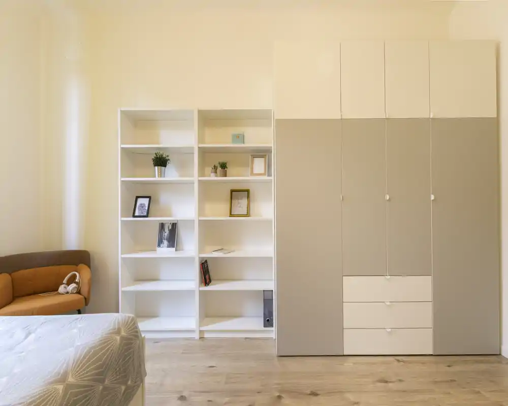 Chambre lumineuse et minimaliste avec coin de lit visible, étagères blanches et grand placard; sol en bois clair et petit canapé orange apportant une touche chaleureuse.