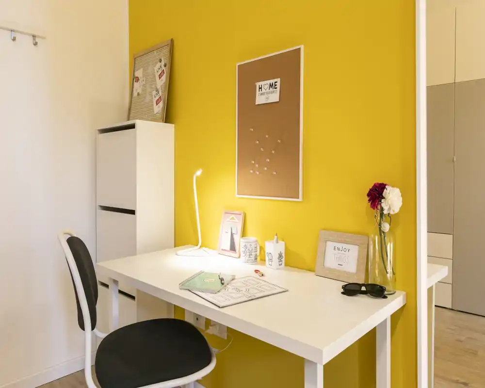 Coin bureau moderne et lumineux avec un bureau blanc devant un mur jaune vif, éclairé par une lampe et décoré d’un tableau en liège et d’objets — idéal pour le travail ou les études.