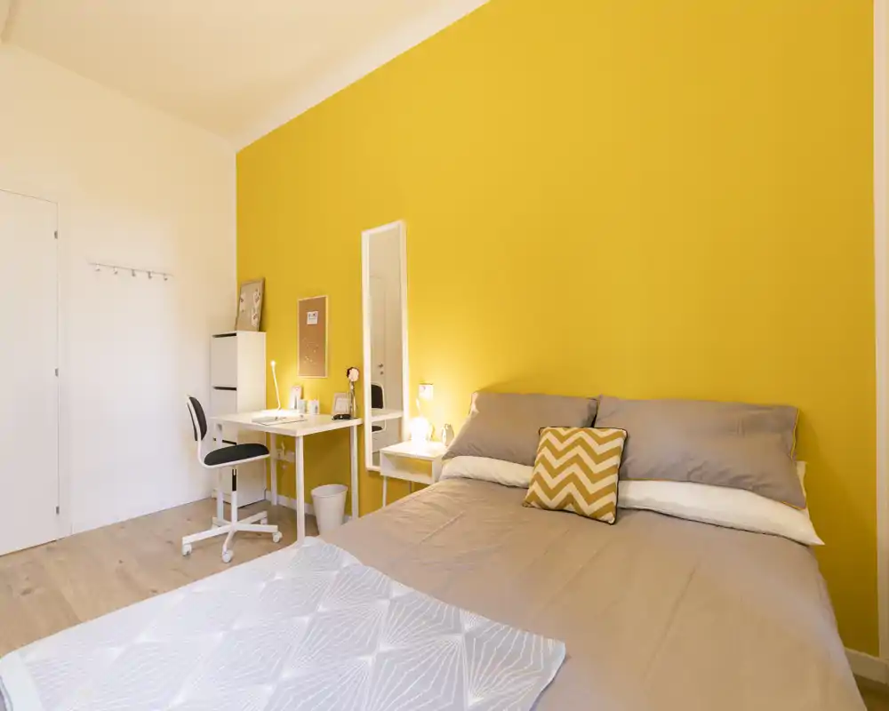 Chambre lumineuse et bien mise en scène avec un mur jaune vif, grand lit aux draps neutres et coussin chevron décoratif ; coin bureau avec bureau et chaise sur le côté.