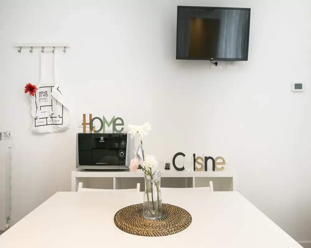 Coin repas minimaliste avec table blanche, dessous de plat tressé et centre de table floral, buffet avec lettres décoratives et télévision murale — lumineux et soigné pour une location courte durée.