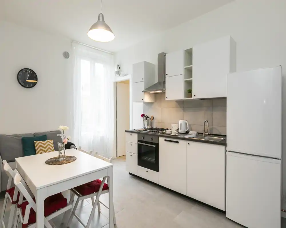 Cuisine ouverte lumineuse avec meubles blancs, four encastré et réfrigérateur, plus une table à manger compacte et un coin salon cosy — idéale pour un petit appartement.