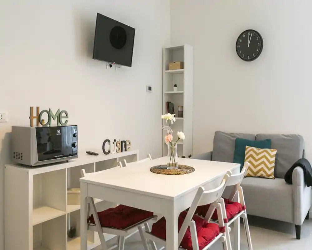 Espace séjour/salle à manger lumineux et soigné avec table blanche, chaises avec coussins rouges, petit canapé et étagères rangées — idéal pour un séjour confortable.