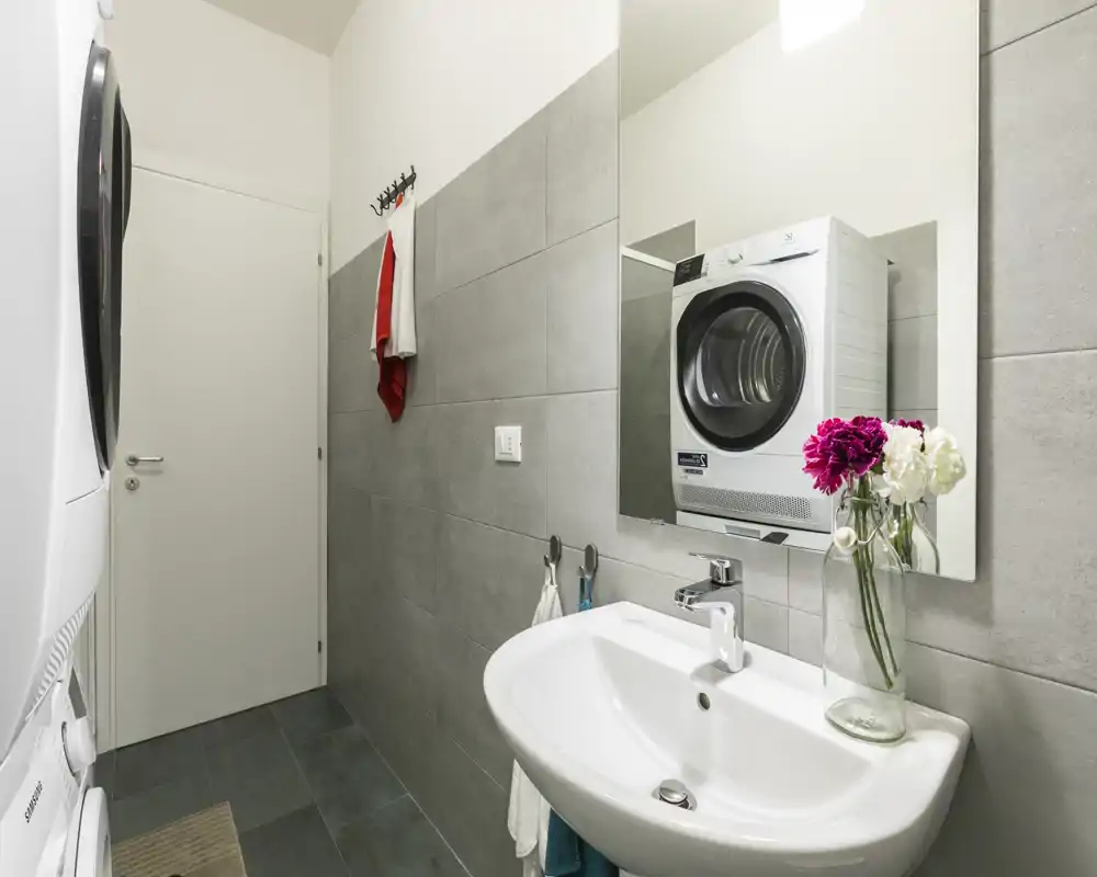 Salle de bains moderne et compacte avec miroir mural, lavabo blanc et machine lavante/séchante empilée visible; carrelage gris et fleurs fraîches offrent un aspect propre et fonctionnel.