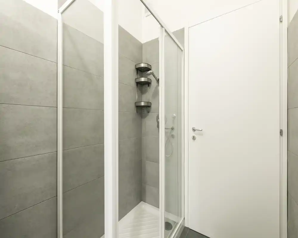 Petite salle de bains moderne avec douche d'angle vitrée, carreaux gris et porte blanche — propre, minimaliste et bien éclairée.