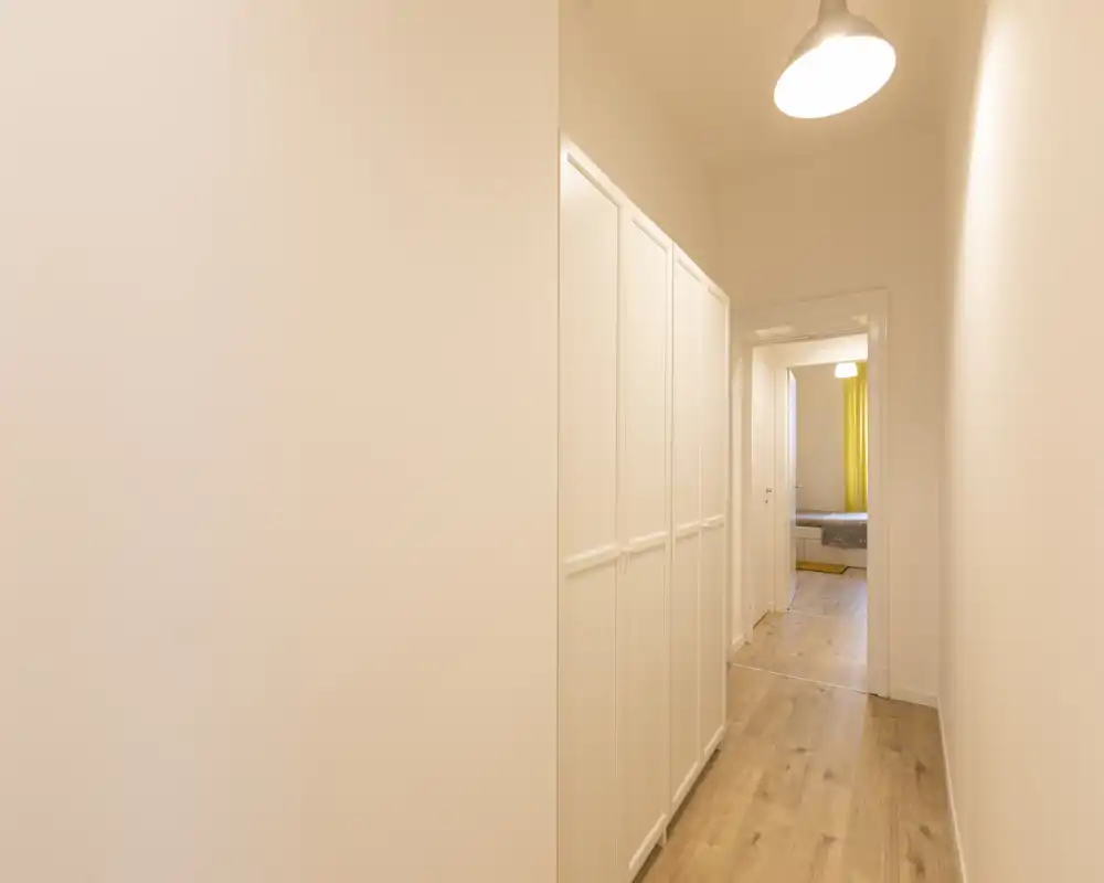 Couloir/entrée étroit(e) bien éclairé(e) avec placards blancs intégrés et sol en bois menant à une chambre ; murs neutres et suspension pour un style propre et minimaliste.