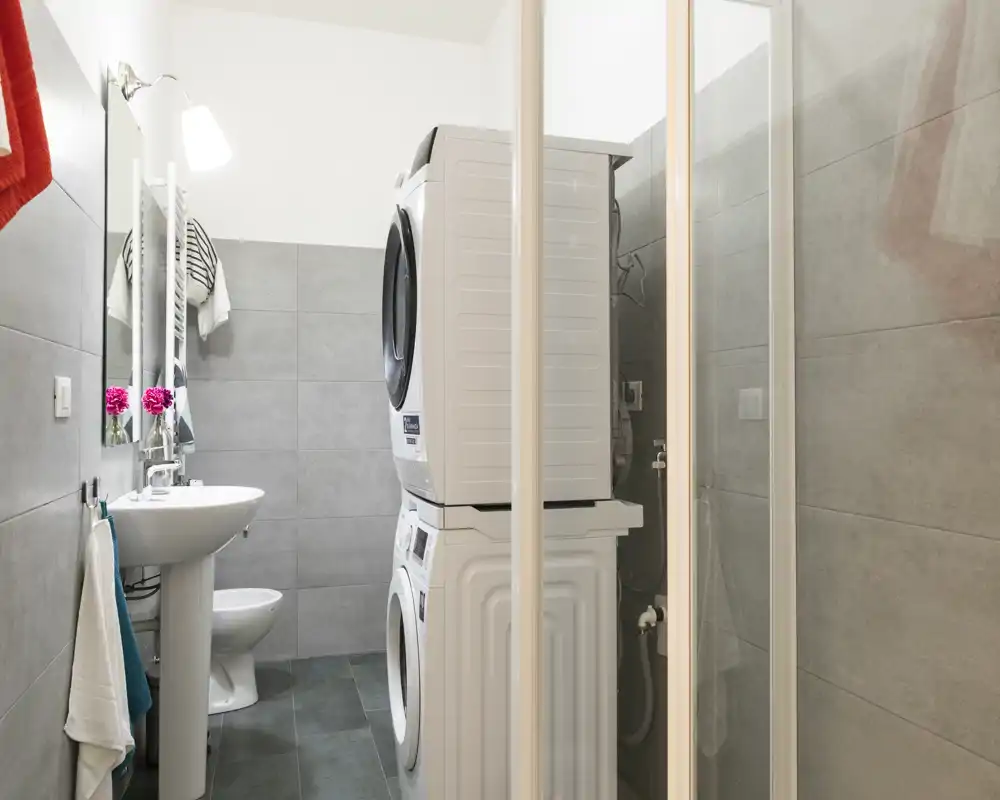 Salle de bain compacte avec murs carrelés gris, lavabo sur pied, WC et une colonne lave-linge/sèche-linge empilée ; agencement moderne et fonctionnel, adapté à la location.