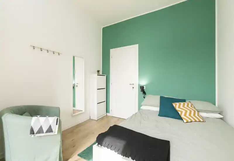 Camera luminosa e curata con letto matrimoniale davanti a una parete verde acqua, cuscini decorativi e una poltrona comoda — ideale per affitti brevi.