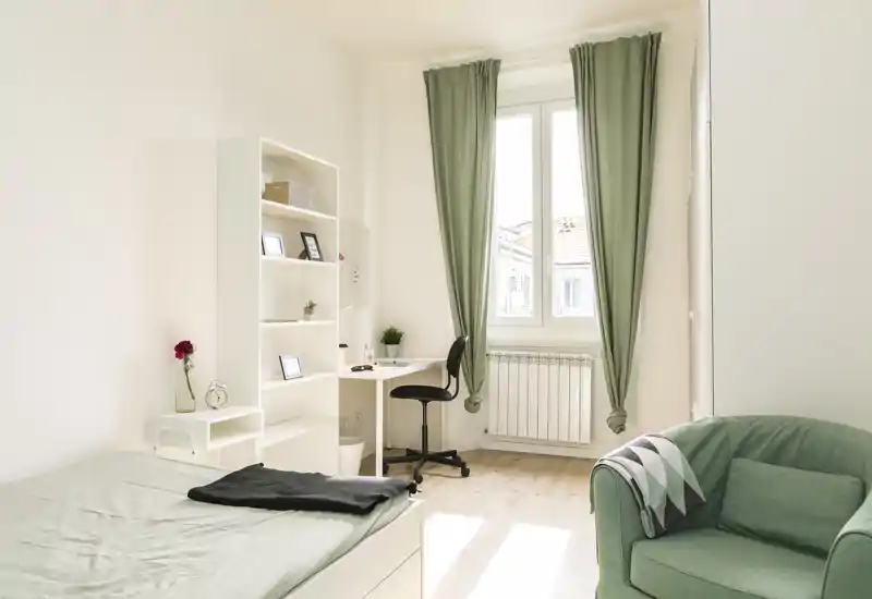 Camera da letto luminosa e minimal con letto singolo, scrivania e poltrona comoda; tende verdi e arredi neutri creano un’atmosfera tranquilla e ariosa.