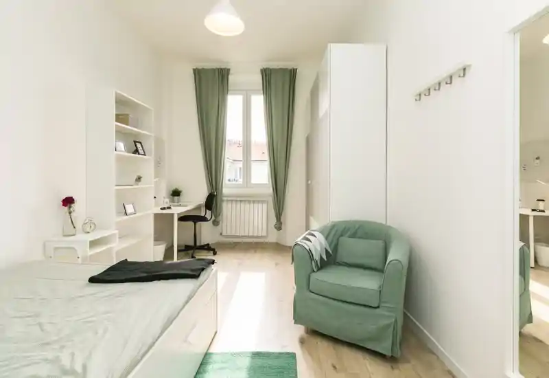 Camera da letto luminosa e ben arredata con letto singolo, poltrona verde, scrivania e armadio; la luce naturale dalla finestra crea un’atmosfera piacevole.