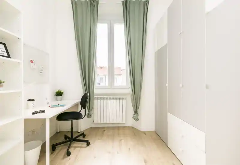 Stanza luminosa e minimal adibita a piccolo spazio living/studio con scrivania, sedia, armadio e una grande finestra con tende verdi.