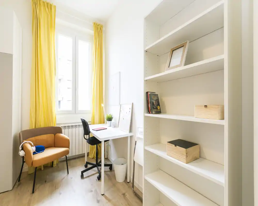 Rincón de estar/estudio luminoso y bien decorado con sillón acogedor, escritorio compacto y estanterías — ideal para teletrabajar o relajarse junto a la ventana.