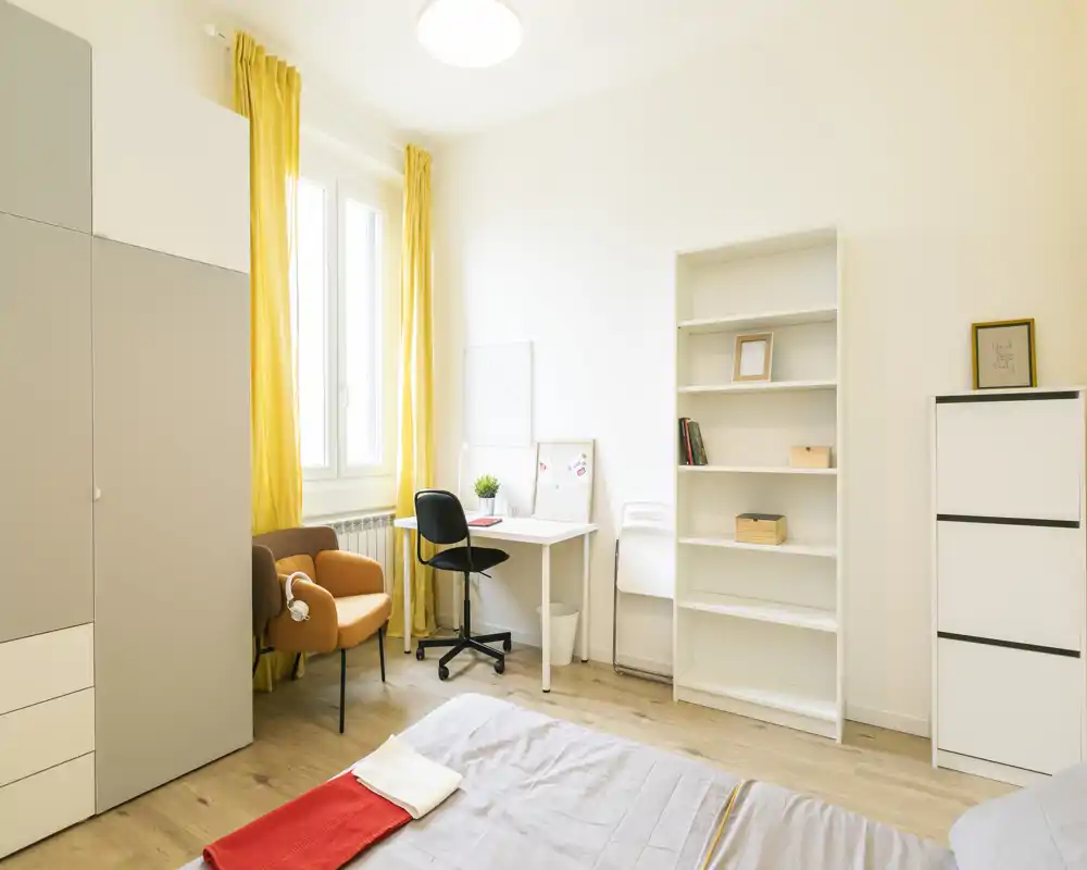 Dormitorio moderno y luminoso con cama arreglada en primer plano, escritorio junto a la ventana, cortinas amarillas y un sillón cómodo—ideal para una persona o estudiante.