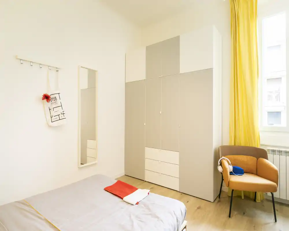 Dormitorio moderno y luminoso con cama bien hecha, gran armario y un sillón acogedor junto a la ventana soleada.