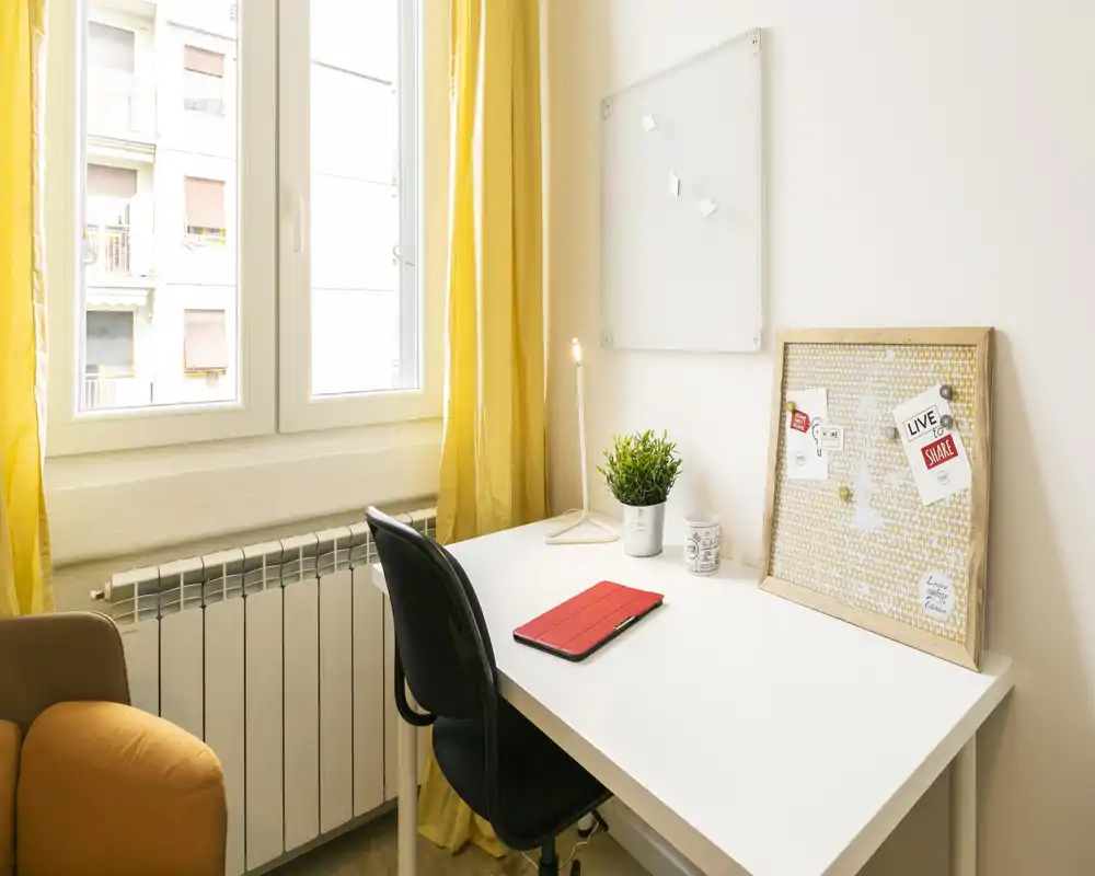 Rincón de oficina luminoso y compacto con mesa blanca, silla ergonómica, tablero de notas y cortinas amarillas; el espacio de trabajo recibe mucha luz natural y está decorado con una planta y una lámpara.