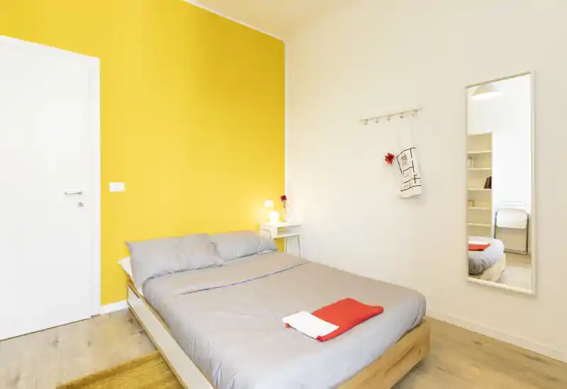 Camera da letto luminosa e moderna con letto matrimoniale, parete accentuata di colore giallo e comodino minimalista. Lenzuola pulite e luce naturale rendono l’ambiente accogliente e perfetto per affitti brevi.