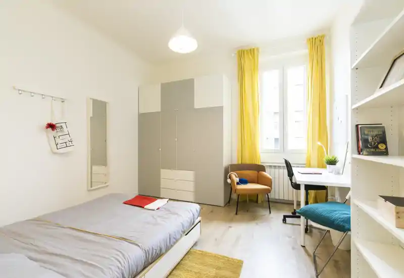 Camera singola luminosa e ben arredata con letto comodo, grande armadio, scrivania e poltrona; ideale per studenti o professionisti. La luce naturale dalla finestra e le tende gialle donano un’atmosfera accogliente.