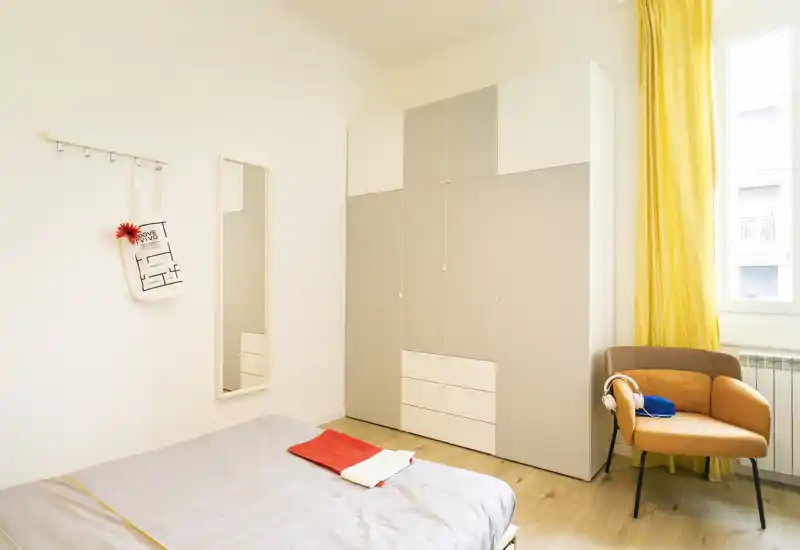 Dormitorio moderno y luminoso con cama bien hecha, gran armario y un sillón acogedor junto a la ventana soleada.