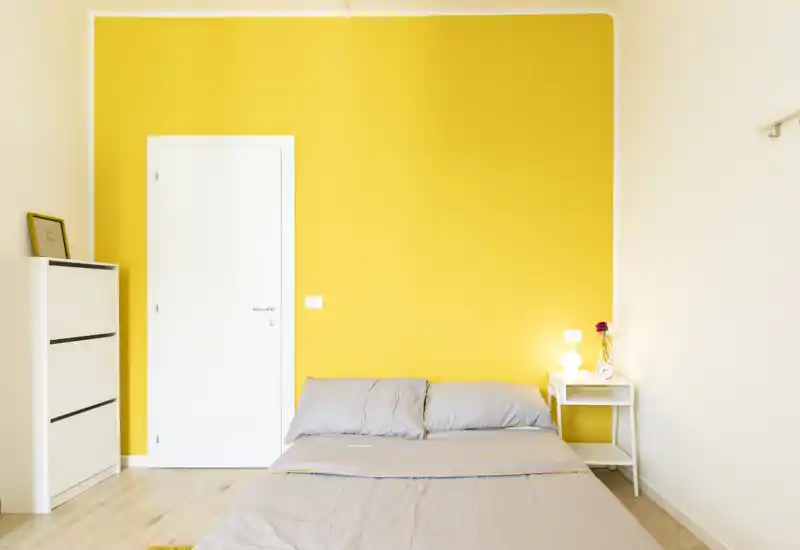 Camera da letto luminosa e moderna con parete gialla vivace, letto matrimoniale ordinato e un comodino minimal con lampada e orologio.
