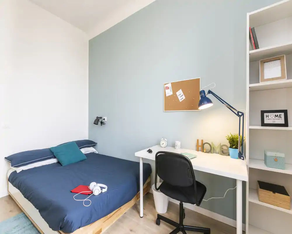 Bright, tidy bedroom with a double bed dressed in navy linens, a small work desk with lamp and shelving—ideal for a single professional or student.