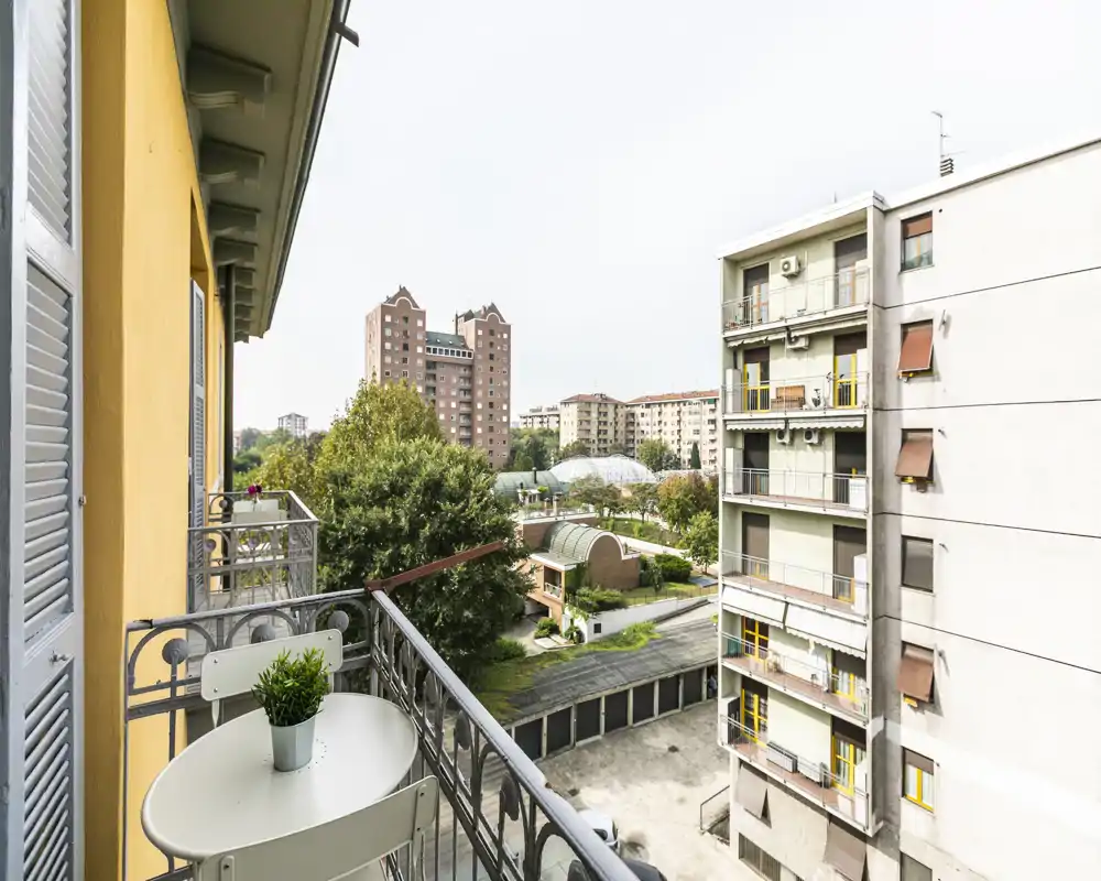Sunny balcony with a small table and chair set, overlooking neighboring buildings and a green courtyard — ideal for morning coffee and city views.