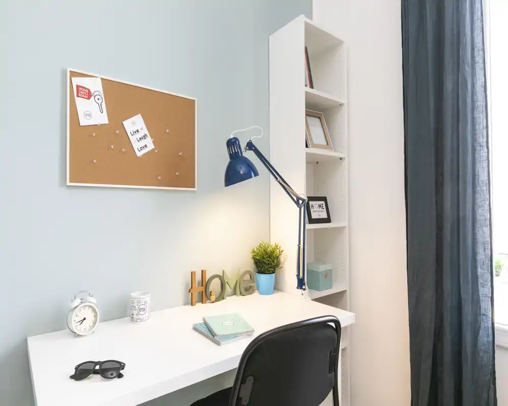A bright, tidy workspace corner with a white desk, blue adjustable lamp, shelving and decorative touches like a small plant and corkboard. Ideal for remote work or study in a calm bedroom setting.