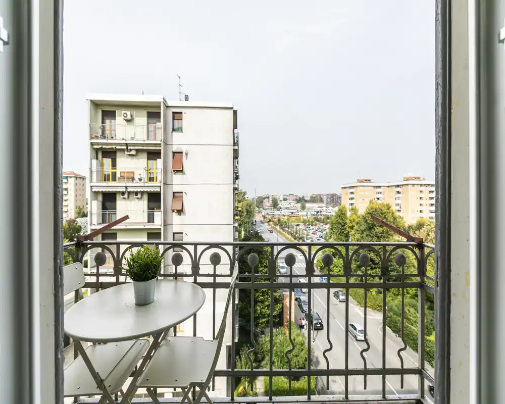 Sunny small balcony with a metal railing, a compact table and two foldable chairs, and a potted plant; view over the street and neighboring apartment blocks.