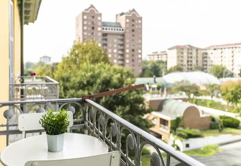 Balcón soleado con una pequeña mesa, dos sillas y una planta en maceta con vistas a un barrio residencial — ideal para el café de la mañana.