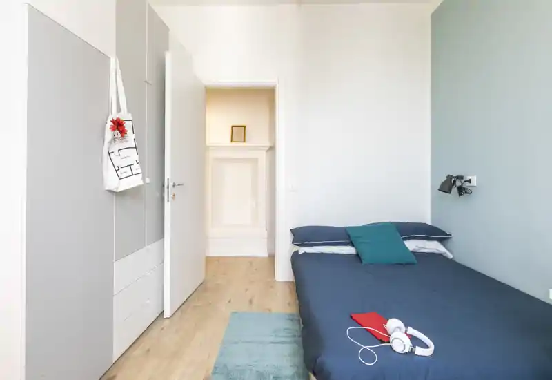 Camera singola moderna e ordinata con letto dal lino blu scuro, lampada da parete, armadio e luce naturale morbida — perfetta come foto per un annuncio di affitto.