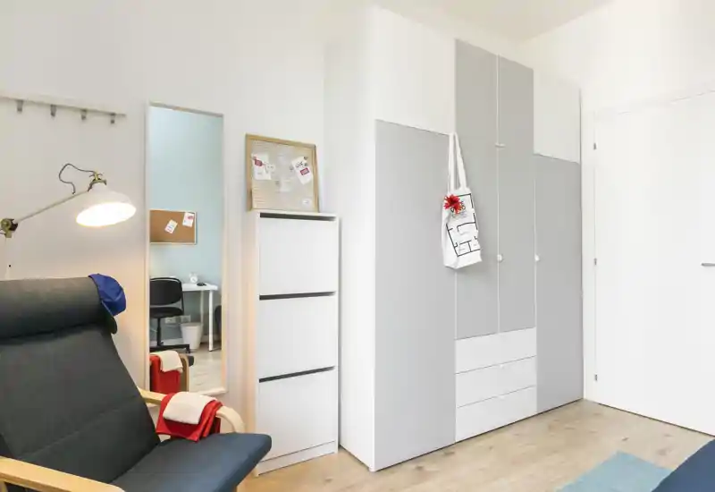 Angolo di camera luminoso e minimale con un ampio armadio, una poltrona comoda e uno specchio intero che riflette una piccola scrivania; stile moderno e pulito ideale per annunci.