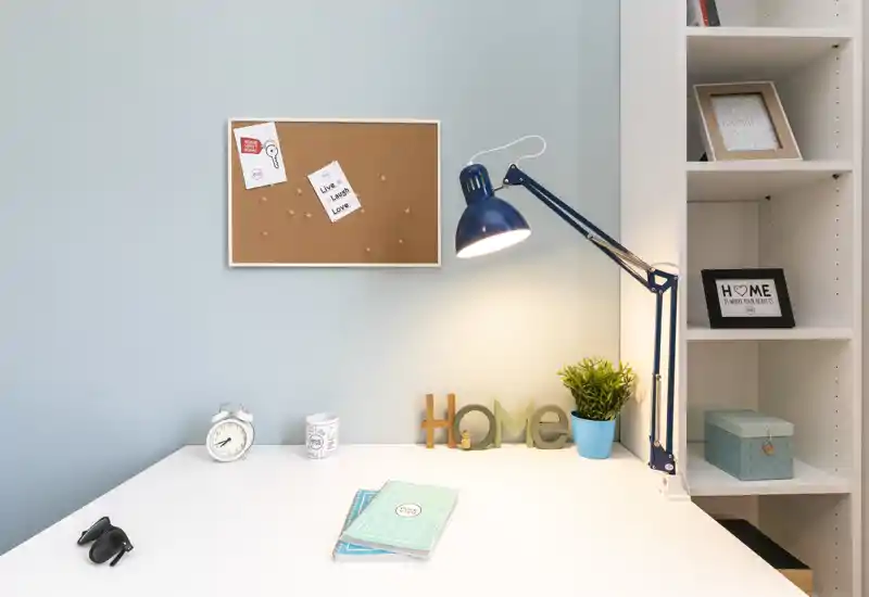 Área de trabajo luminosa y ordenada con escritorio blanco, lámpara orientable, corcho y detalles decorativos como una planta pequeña y las letras 'HOME'.