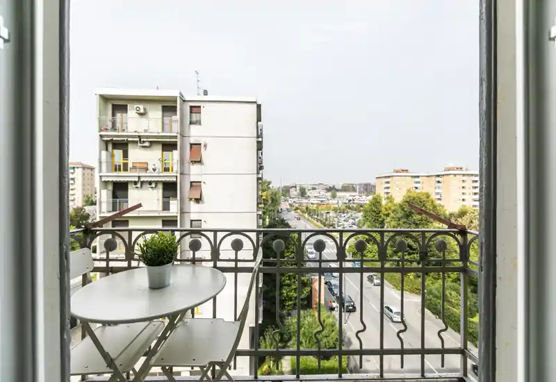 Pequeño balcón soleado con barandilla metálica, mesa compacta y dos sillas plegables y una planta en maceta; vistas a la calle y a los bloques de apartamentos cercanos.