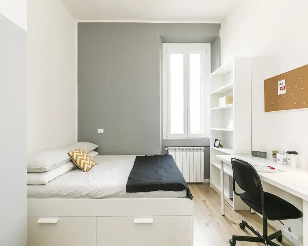 Dormitorio luminoso y minimalista con cama doble, almacenamiento bajo la cama, escritorio y estanterías junto a la ventana — ideal para estudiantes o profesionales.