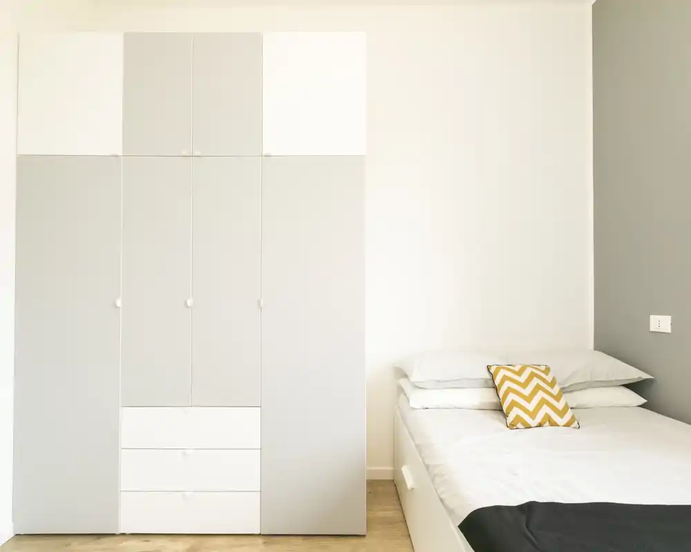 Dormitorio luminoso y minimalista con cama individual y armario empotrado; tonos neutros y cojín con estampado zigzag aportan un aire moderno y sereno — ideal para anuncios que destaquen almacenamiento y sencillez.