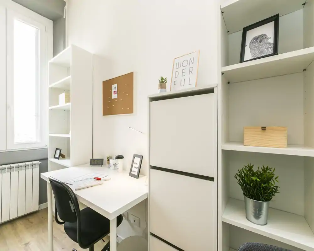 Pequeño espacio de trabajo moderno y luminoso con escritorio blanco, estanterías y luz natural de una gran ventana — ideal para estudiar o trabajar desde casa.