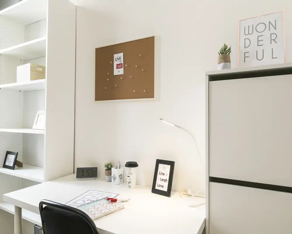 Área de trabajo en casa luminosa con escritorio blanco, estantería y lámpara — ideal para estudiar o teletrabajar. La imagen destaca una mesa ordenada con pequeñas plantas y marcos decorativos.