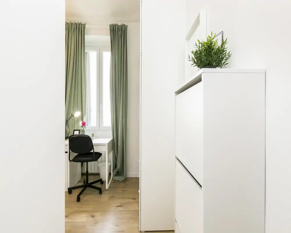 Pequeño espacio de trabajo/entrada luminoso con escritorio junto a la ventana, cortinas verdes y un mueble blanco. Ambiente limpio que destaca la luz natural y un estilo nórdico.