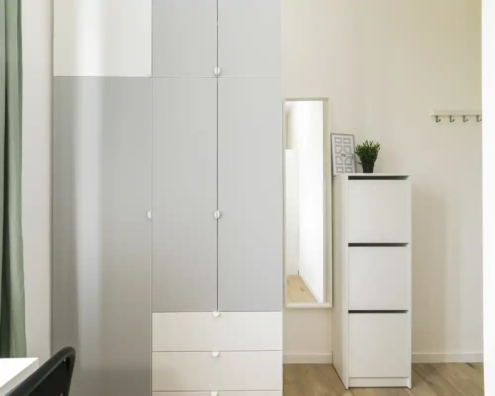 Zona de dormitorio minimalista y luminosa centrada en el almacenamiento: un armario alto, una cómoda estrecha y un espejo de cuerpo entero sobre suelo de madera cálido.