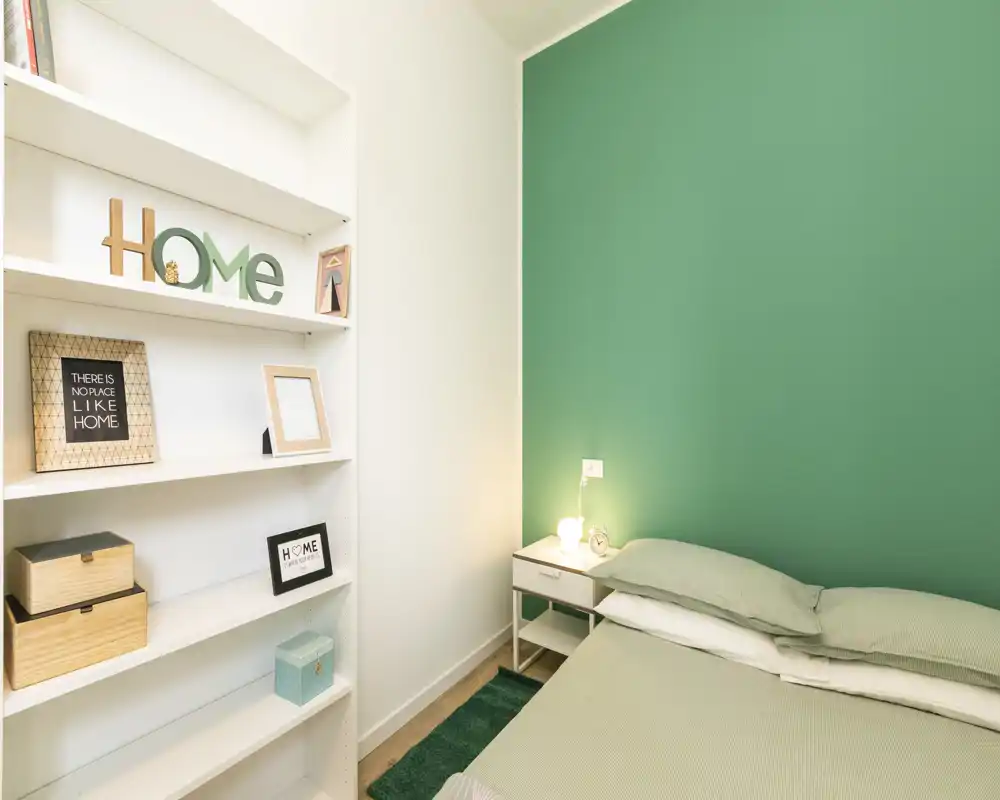 Dormitorio acogedor y bien iluminado con pared verde, mesita de noche y lámpara. La cama y los textiles son claramente visibles, y una estantería empotrada con objetos decorativos añade personalidad.