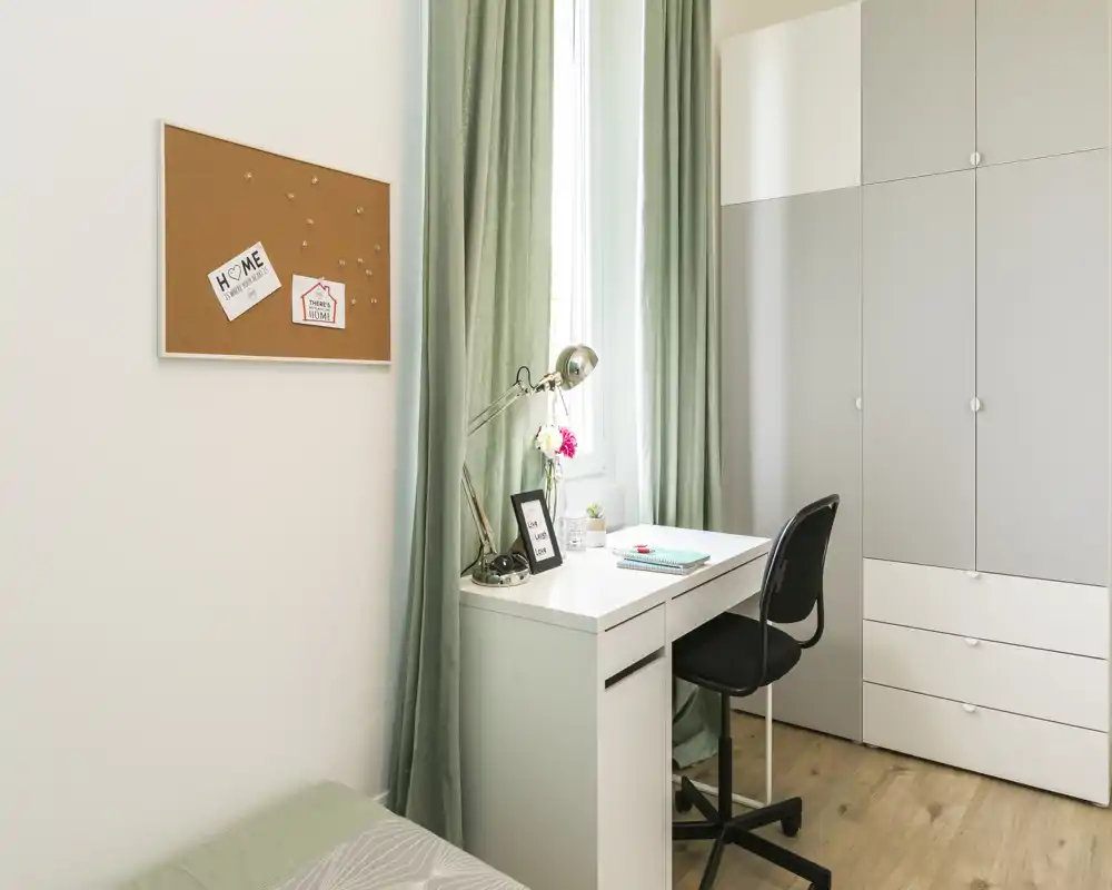 Rincón de dormitorio luminoso y ordenado con un escritorio blanco, lámpara de trabajo y armario. Cortinas verde suave y un tablero de corcho crean un área de trabajo funcional junto a la cama.