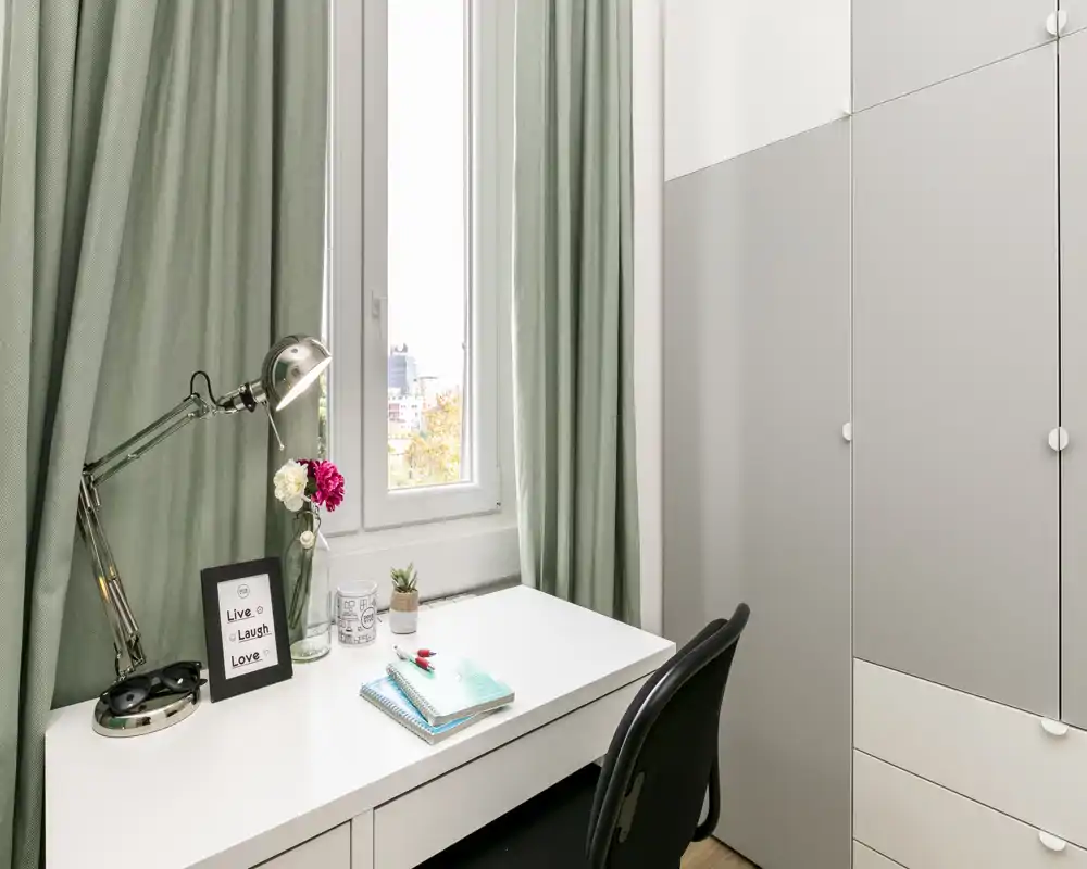 Rincón luminoso y ordenado con un escritorio blanco, lámpara de trabajo, silla y armario empotrado junto a una ventana; adecuado como zona de estudio en el dormitorio.