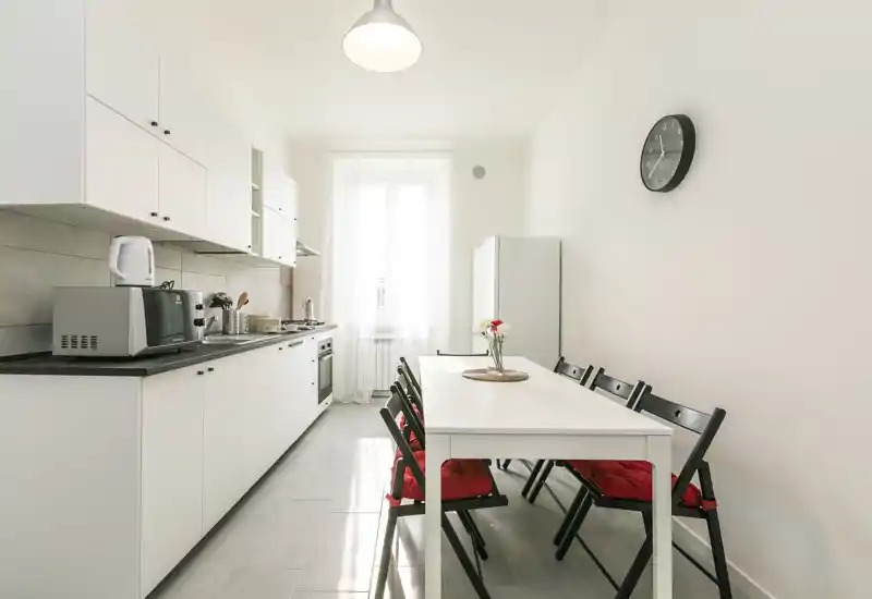 Cocina luminosa y minimalista con una larga encimera, horno empotrado, microondas y mesa de comedor con cuatro sillas; muebles blancos y luz natural desde la ventana que crean una sensación fresca y moderna.