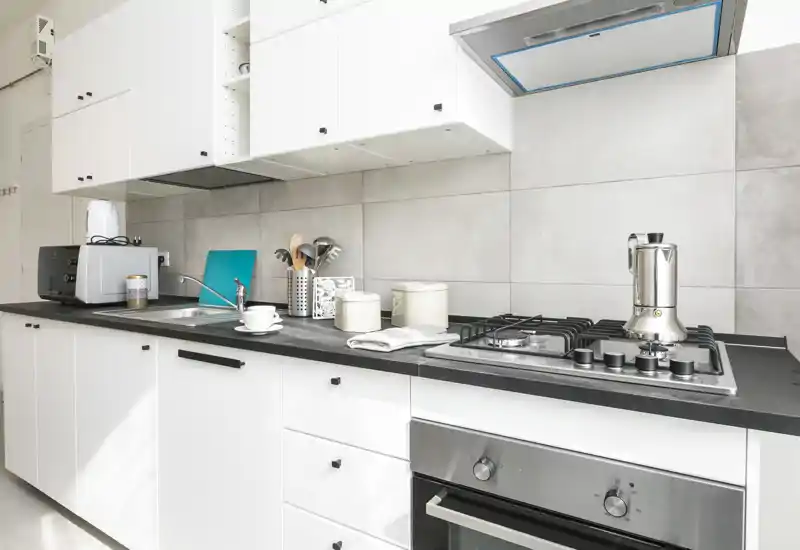Cocina moderna blanca con horno empotrado, placa de gas y cafetera moka de acero, encimeras limpias y armarios prácticos.