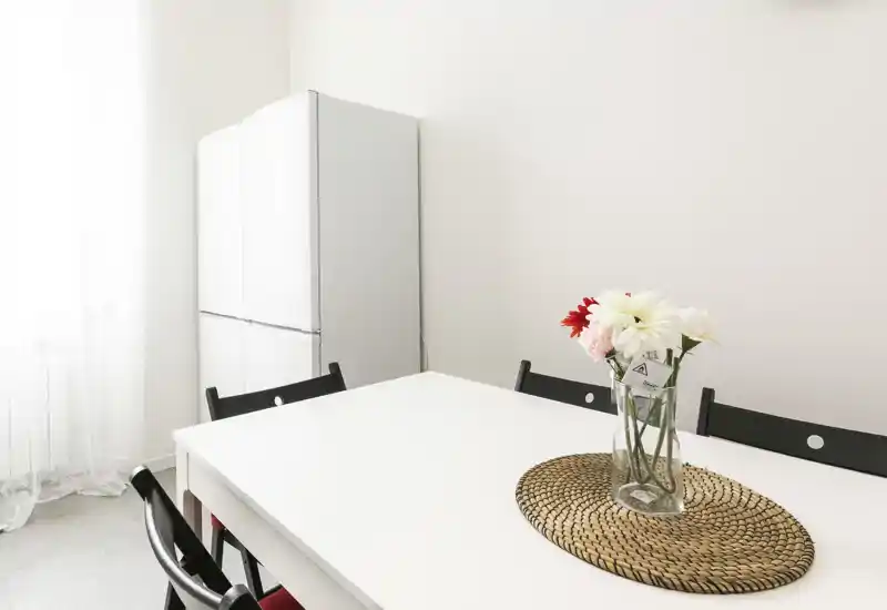 Zona de comedor luminosa y minimalista con mesa blanca, sillas negras y un jarrón con flores sobre un salvamantel trenzado; la luz natural realza la limpieza del espacio.