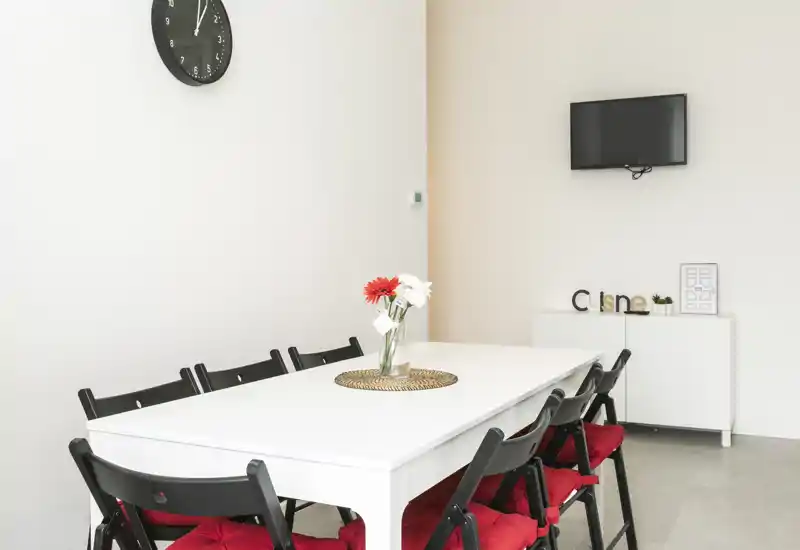Zona de comedor luminosa y minimalista con mesa blanca, seis sillas negras con cojines rojos y un jarrón decorativo — perfecta para comidas compartidas y anuncios de pisos compactos.