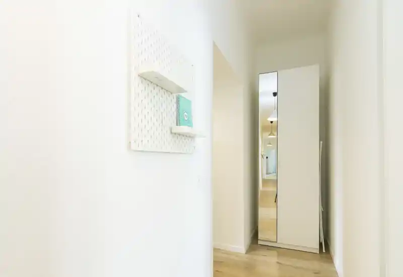 Entrada luminosa y minimalista con suelo de madera clara, un armario alto con espejo y estantes de pared sencillos — espacio de acceso moderno y limpio.