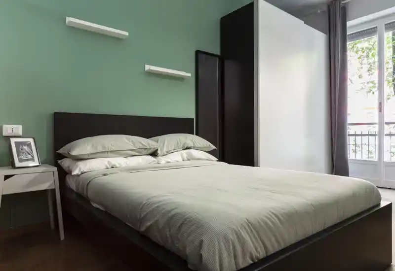 Bright, modern bedroom with a large double bed, soft linens and natural light from a balcony window. Simple, elegant decor with a mint-green accent wall and practical wardrobe.
